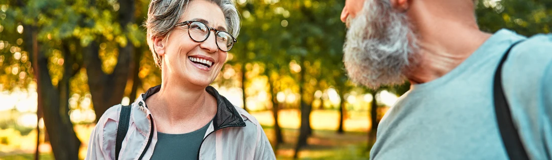 Two people standing outdoors in a park-like setting with green trees in the background, engaged in conversation. Both are wearing casual clothing and carrying shoulder bags.