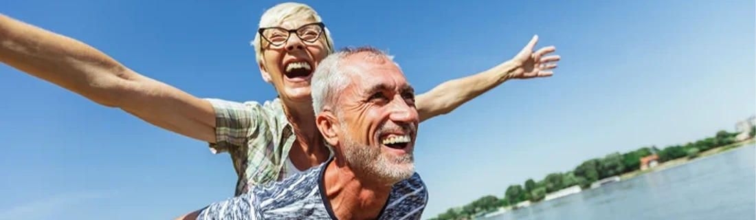 Two people enjoying a playful moment near a body of water, with one person leaning forward and the other extending arms wide as if mimicking an airplane. The sky is clear and blue, and there are trees and buildings in the distance.